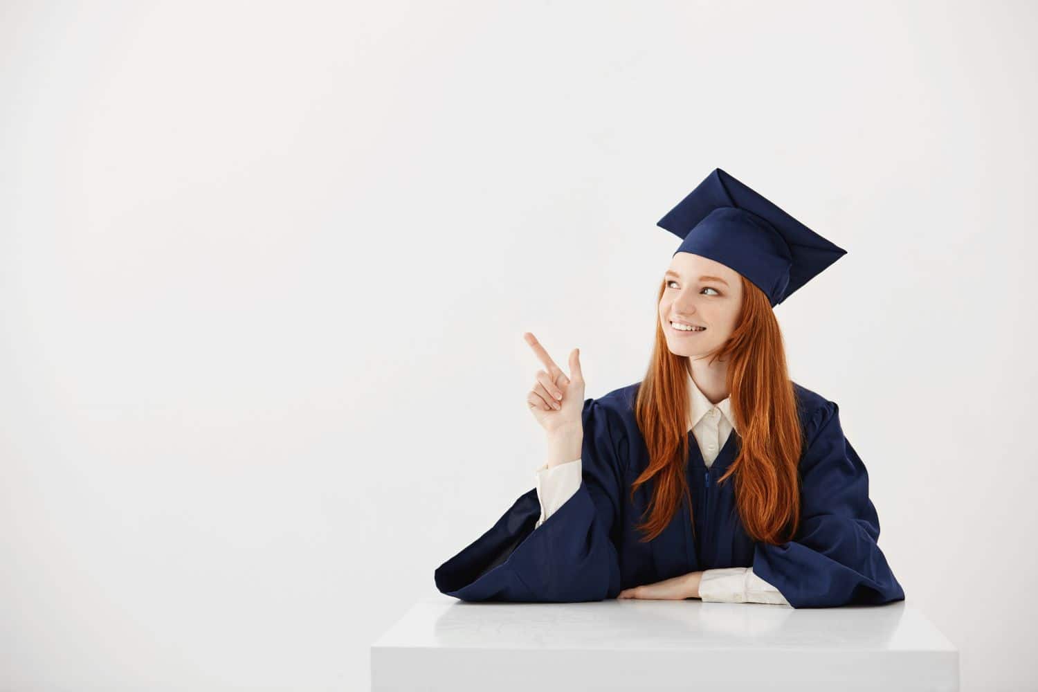 young-female-university-graduate-academic-cap-sitting-table-smiling-pointing-left-future-lawyer-engineer-showing-idea1